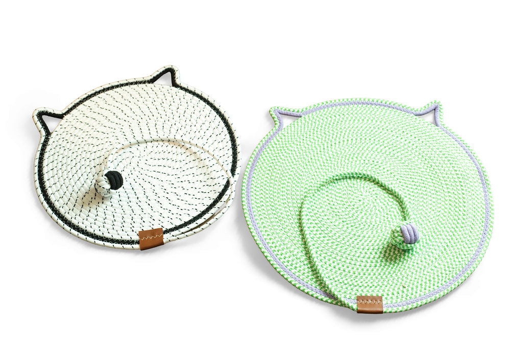 Two round woven pet mats with cat-ear cutouts, shown on a white background. One mat is off-white with black trim and a black handle, and the other is light green with white trim and a purple handle.